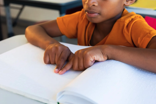 A child in an orange shirt reads a Braille book by running their fingers over the raised dots on the page.
